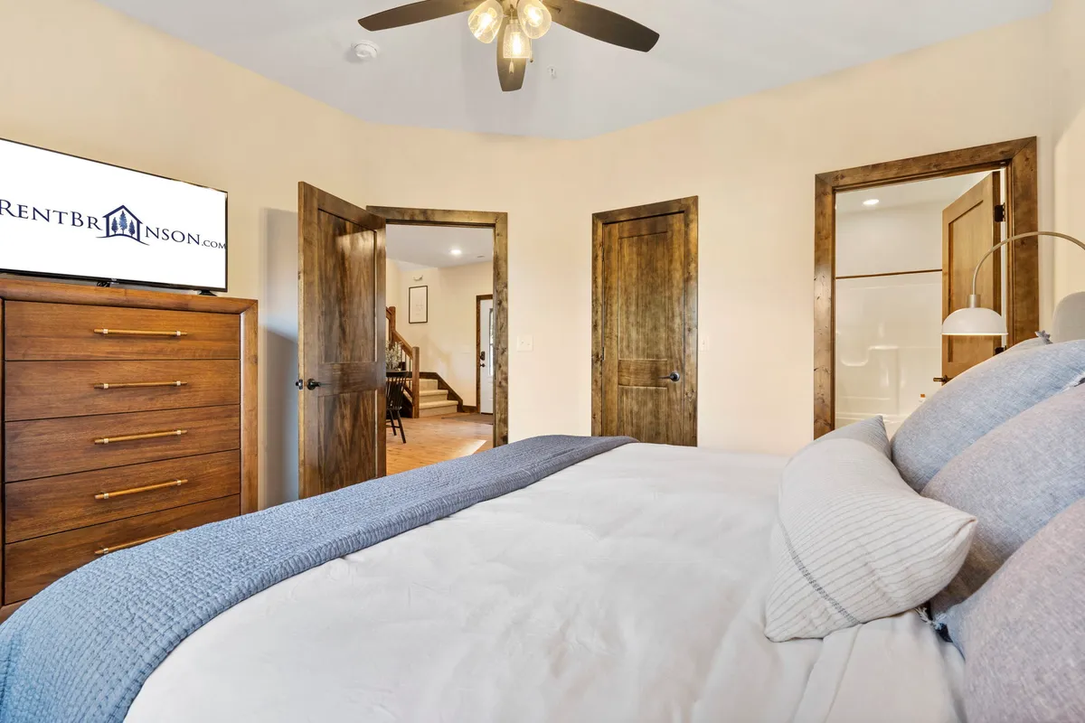 A cozy king bedroom featuring decorative pillows, a wooden dresser, a television, and doorways to other rooms, with soft morning sunlight streaming in.