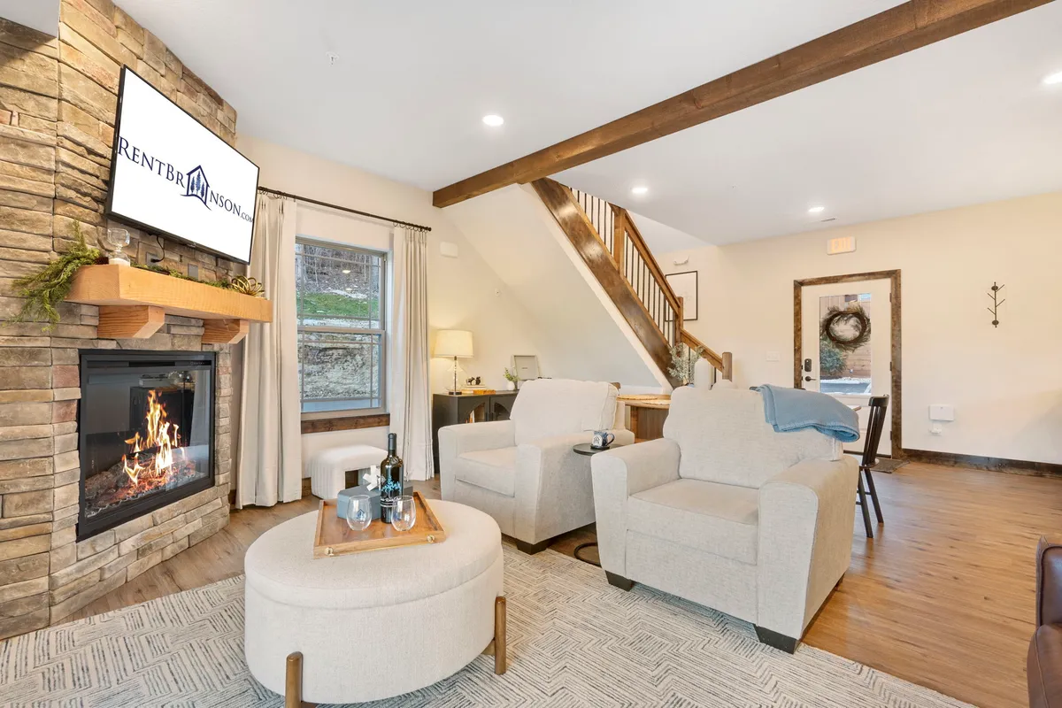The image showcases a warm, inviting cabin interior with a crackling stone fireplace as the focal point. Comfortable chairs are arranged around a rustic coffee table, with a TV nearby and a wooden staircase leading to the upper level. The space exudes coziness and charm, highlighted by wood accents and soft lighting.