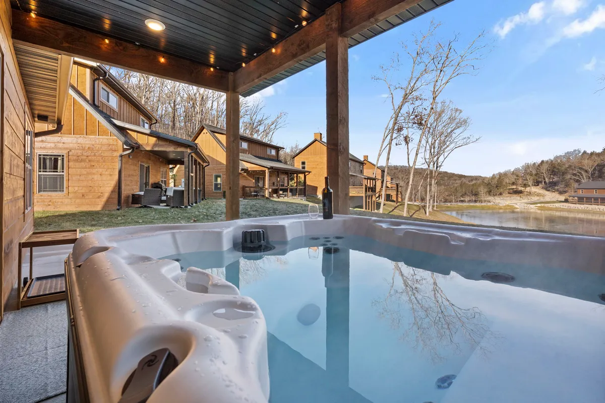 This image depicts an inviting hot tub situated outdoors with wooden cabins in the background and a stunning lake view. The setting is serene, with people relaxing in the hot tub and others enjoying the tranquil lakeside ambiance. The lighting suggests either an early morning or sunset, highlighting the peaceful atmosphere.