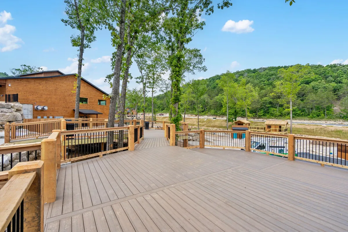 A wooden deck surrounded by tall trees with distant hills visible in the background. The deck is open, inviting, and offers an unobstructed view into the lush nature beyond. Sunlight filters gently through the foliage, enhancing the tranquil atmosphere.