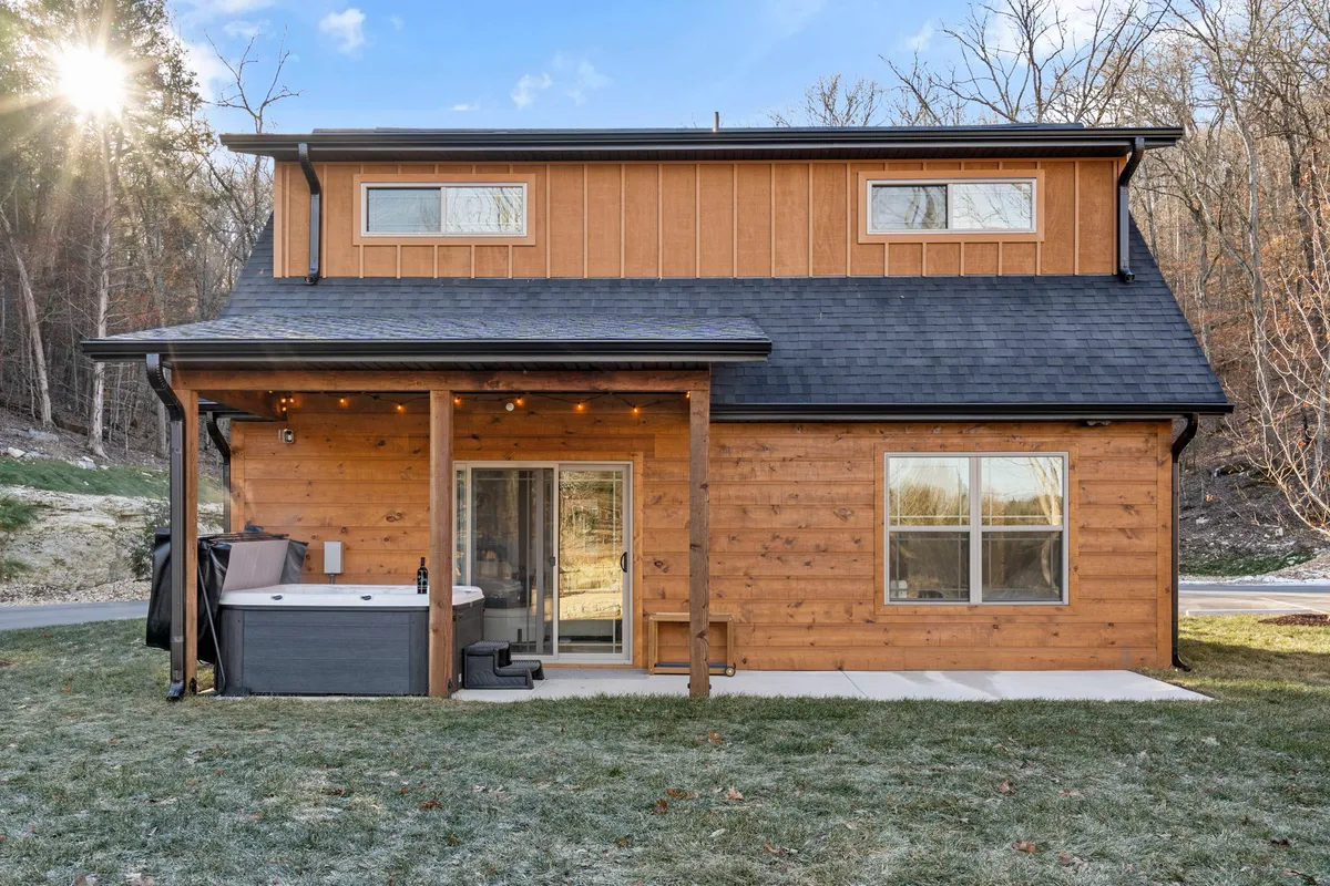 This image displays the cozy wooden exterior of Clearview Cottage, featuring large windows and a welcoming patio complete with a hot tub. The patio area is inviting, surrounded by nature, and designed for relaxation. The image is captured during sunset, bathing the space in a warm and romantic glow.