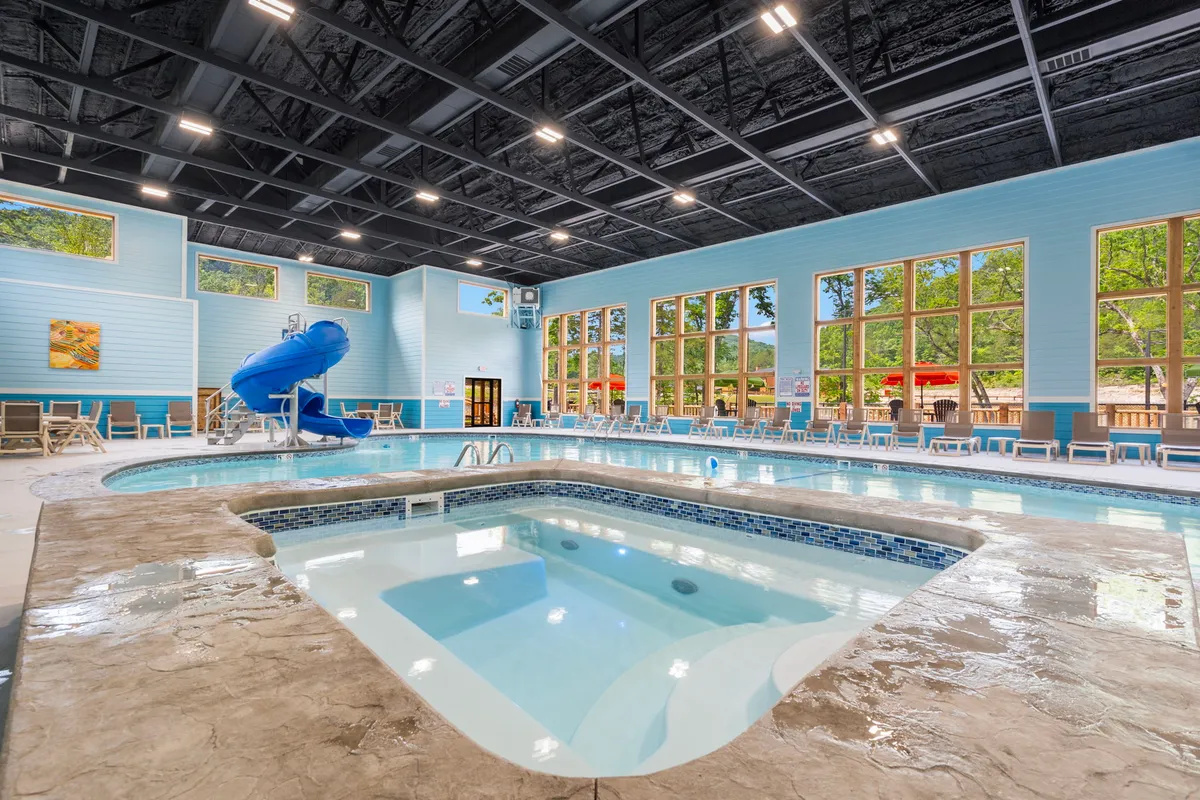 An indoor swimming pool area featuring a water slide, jacuzzi, large windows letting in natural light, and ample poolside seating. Children are seen playing on the slide, while families relax by the water or in the jacuzzi, creating a lively and inviting atmosphere.