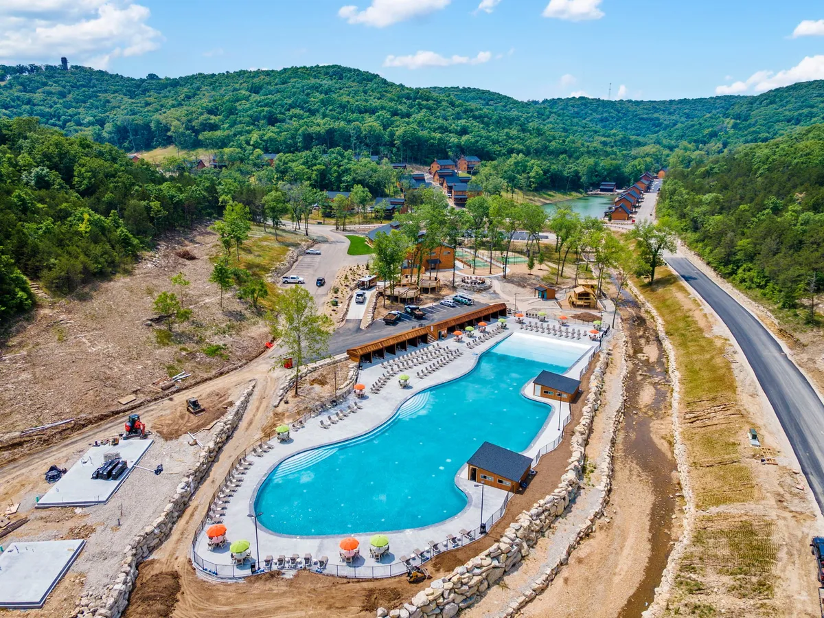 A spacious outdoor swimming pool is surrounded by lush greenery and a lodge-style building. Deck chairs line the poolside, inviting guests to relax, swim, and enjoy sunny afternoons with family and friends.