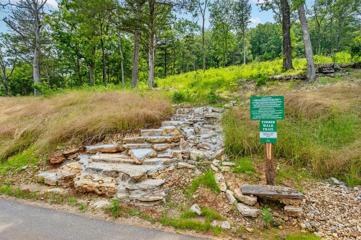 This image shows a peaceful forest trail bordered by stone steps and lush greenery, with a trail sign inviting visitors to explore the natural surroundings. The dense trees and vibrant plant life evoke a sense of immersion in nature and hint at opportunities for hiking, birdwatching, and quiet reflection.