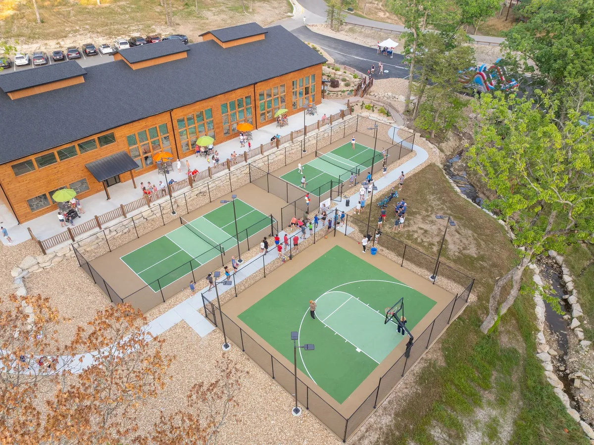 A lively gathering unfolds outside a large wooden building, with tennis and basketball courts active in the foreground. Groups of people are socializing, playing sports, and enjoying the festive, community atmosphere under open skies.