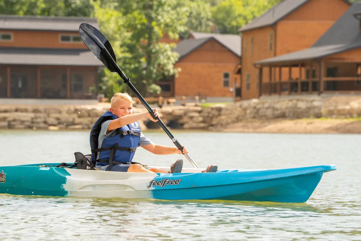 A young boy paddles a kayak across the calm surface of Table Rock Lake, with charming wooden cabins dotting the shoreline in the background. The scene is bright and energetic, capturing the joy of summer days spent on the water, with a clear connection to lakeside activities and family fun.