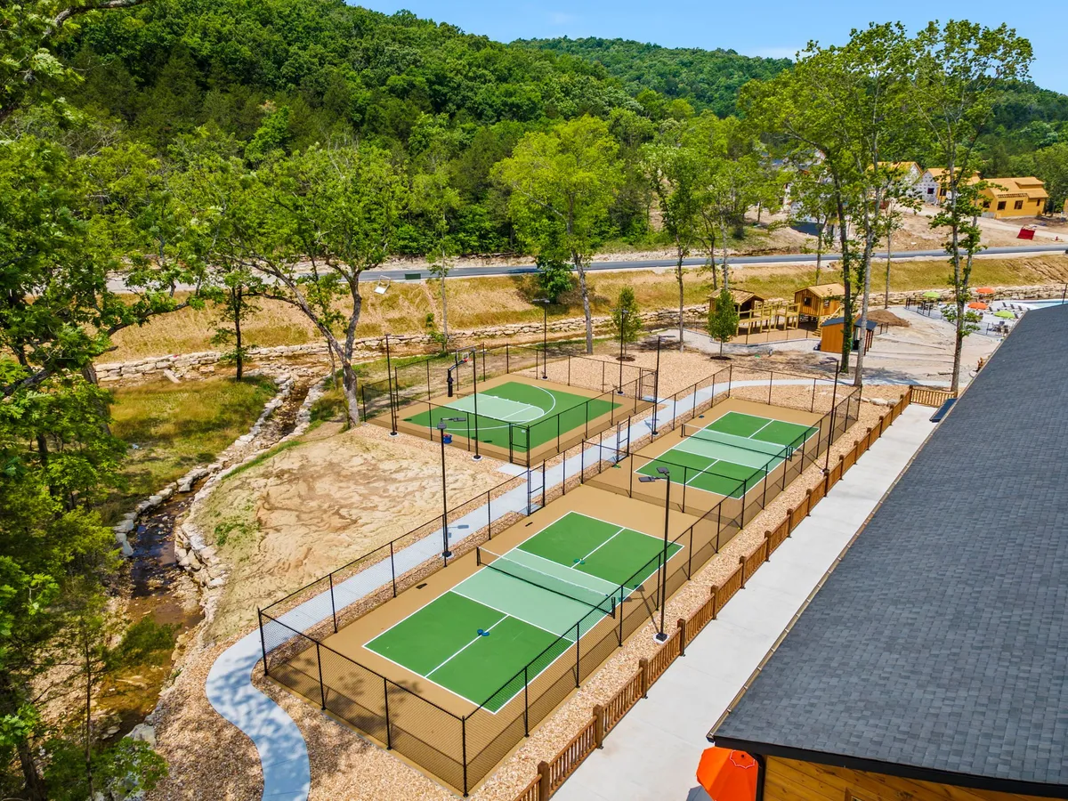 Tennis courts set amid trees, with a creek and a playground nearby, creating a vibrant area for sports and family outings. The mood is energetic and playful, with facilities for tennis and play.