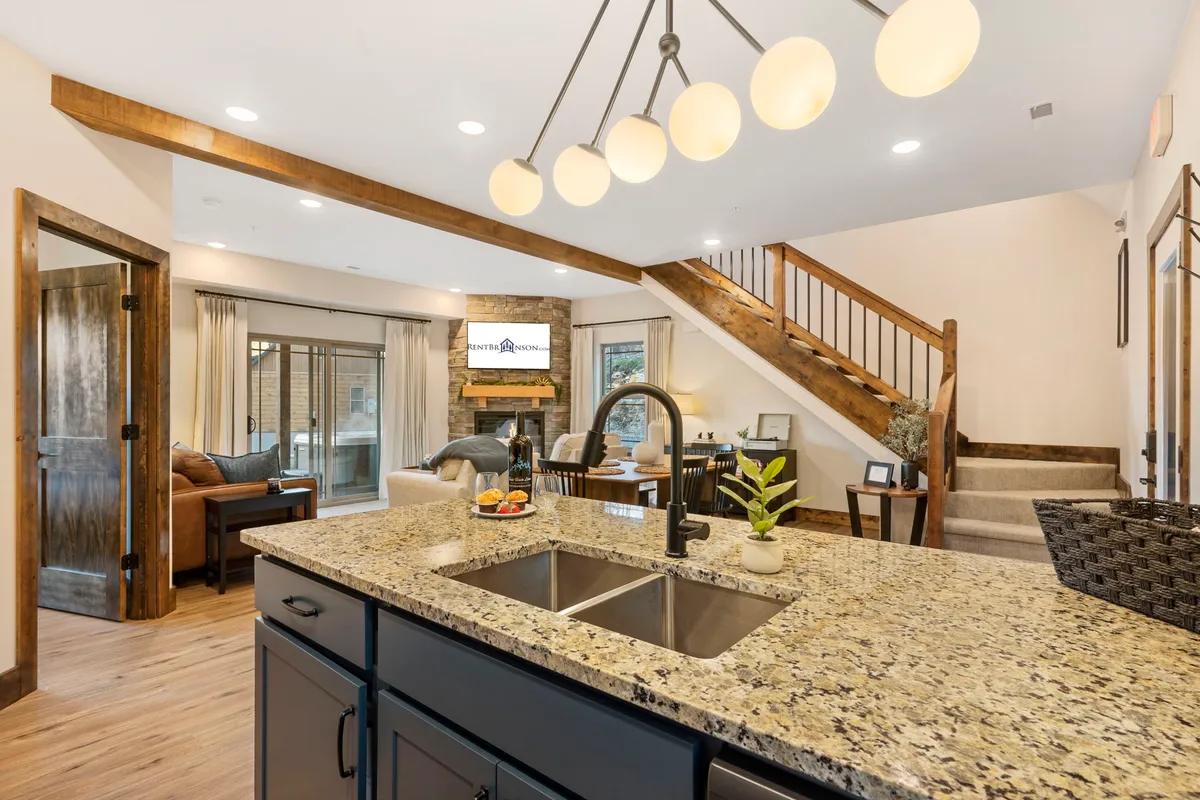 An inviting, open-concept interior featuring a central kitchen island with granite countertop, chic pendant lighting, a stone fireplace, comfortable seating area, and a staircase leading to an upper level. Large windows bring in abundant natural light, highlighting the seamless transition between the living, dining, and kitchen spaces.