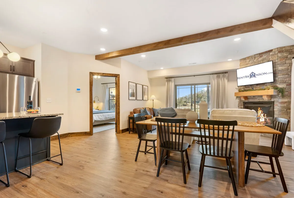 A warm and stylish interior scene showing a dining table set beside a modern fireplace and a kitchen island, all framed by large windows that offer serene wooded vistas. The layout encourages shared meals, game nights, and relaxed evenings by the fire.